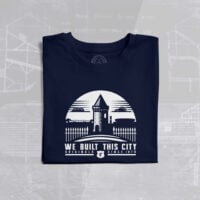 The image shows a folded navy blue T-shirt with a white graphic design on the front. The design features a stylized illustration of Goodison Park, Everton Football Club's historic stadium, with its iconic Archibald Leitch latticework and a prominent tower structure. Above the stadium illustration, there is a semi-circle with a triangular shape at the top, resembling a rising sun or a badge. Below the illustration, the text reads: 