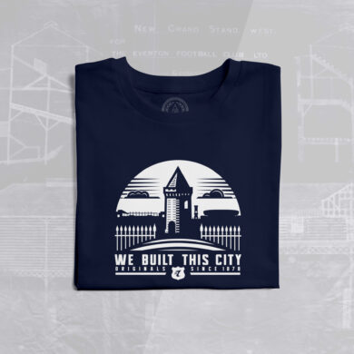 The image shows a folded navy blue T-shirt with a white graphic design on the front. The design features a stylized illustration of Goodison Park, Everton Football Club's historic stadium, with its iconic Archibald Leitch latticework and a prominent tower structure. Above the stadium illustration, there is a semi-circle with a triangular shape at the top, resembling a rising sun or a badge. Below the illustration, the text reads: "WE BUILT THIS CITY ORIGINALS SINCE 1878." The word "ORIGINALS" is in bold, and "SINCE 1878" is smaller, with a small Everton FC crest (a shield with the initials "EFC" and a tower) placed between the two phrases. In the background, there is a faint blueprint or architectural drawing of Goodison Park, with text such as "NEW GRAND STAND WEST" and "THE EVERTON FOOTBALL CLUB LTD" visible, adding a historical and nostalgic touch to the design. The T-shirt is laid flat on a light gray surface.