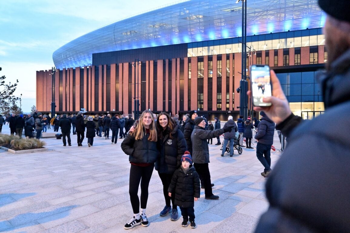 A group of Everton fans posing for a photo outside Everton's new stadium, a modern structure with a glass and wood-paneled exterior. The stadium is lit up with blue lights, and a crowd of people in winter jackets gathers in the foreground. Two children and an adult stand together, smiling, with one child wearing a blue and red beanie. Another person in the foreground takes a photo with a smartphone, capturing the lively atmosphere before a match.