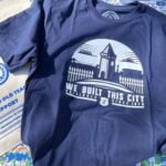 A navy blue T-shirt and a beige tote bag are laid out on a colorful background featuring cartoon illustrations of Everton Football Club fans, players, and banners. The T-shirt has a white graphic design showing a silhouette of Goodison Park's iconic tower and fence, with the text "WE BUILT THIS CITY ORIGINALS SINCE 1878" below it. The tote bag has a blue circular logo with a tower and the text "GOT" in the center, surrounded by a wreath. Below the logo, the bag reads "IT'S A GRAND OLD TEAM TO SUPPORT FOR EVERTONIANS, BY EVERTONIANS WWW.GRANDOLDTEAM.COM" in blue text, with a smaller note at the bottom saying "PLEASE KEEP THIS BAG OUT OF REACH OF BABIES AND SMALL CHILDREN." The background includes various Everton-related slogans and imagery, such as "EVERTON F.C. SINCE 1878" and "GOODISON PARK."