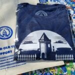 A navy blue T-shirt and a beige tote bag are laid out on a colorful background featuring cartoon illustrations of Everton Football Club fans, players, and banners. The T-shirt has a white graphic design showing a silhouette of Goodison Park's iconic tower and fence, with the text "WE BUILT THIS CITY ORIGINALS SINCE 1878" below it. The tote bag has a blue circular logo with a tower and the text "GOT" in the center, surrounded by a wreath. Below the logo, the bag reads "IT'S A GRAND OLD TEAM TO SUPPORT FOR EVERTONIANS, BY EVERTONIANS WWW.GRANDOLDTEAM.COM" in blue text, with a smaller note at the bottom saying "PLEASE KEEP THIS BAG OUT OF REACH OF BABIES AND SMALL CHILDREN." The background includes various Everton-related slogans and imagery, such as "EVERTON F.C. SINCE 1878" and "GOODISON PARK."
