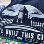 A navy blue T-shirt and a beige tote bag are laid out on a colorful background featuring cartoon illustrations of Everton Football Club fans, players, and banners. The T-shirt has a white graphic design showing a silhouette of Goodison Park's iconic tower and fence, with the text "WE BUILT THIS CITY ORIGINALS SINCE 1878" below it. The tote bag has a blue circular logo with a tower and the text "GOT" in the center, surrounded by a wreath. Below the logo, the bag reads "IT'S A GRAND OLD TEAM TO SUPPORT FOR EVERTONIANS, BY EVERTONIANS WWW.GRANDOLDTEAM.COM" in blue text, with a smaller note at the bottom saying "PLEASE KEEP THIS BAG OUT OF REACH OF BABIES AND SMALL CHILDREN." The background includes various Everton-related slogans and imagery, such as "EVERTON F.C. SINCE 1878" and "GOODISON PARK."