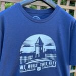 A navy blue T-shirt is hung on a hanger against a wooden lattice fence with greenery in the background. The T-shirt features a white graphic design with a silhouette of Goodison Park's tower and fence, set against a circular background with clouds. Below the graphic, the text reads "WE BUILT THIS CITY ORIGINALS SINCE 1878" in white, with a small Everton logo in the center of the text. The T-shirt has a casual, short-sleeve design.