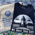 A navy blue T-shirt and a beige tote bag are laid out on a colorful background featuring cartoon illustrations of Everton Football Club fans, players, and banners. The T-shirt has a white graphic design showing a silhouette of Goodison Park's iconic tower and fence, with the text "WE BUILT THIS CITY ORIGINALS SINCE 1878" below it. The tote bag has a blue circular logo with a tower and the text "GOT" in the center, surrounded by a wreath. Below the logo, the bag reads "IT'S A GRAND OLD TEAM TO SUPPORT FOR EVERTONIANS, BY EVERTONIANS WWW.GRANDOLDTEAM.COM" in blue text, with a smaller note at the bottom saying "PLEASE KEEP THIS BAG OUT OF REACH OF BABIES AND SMALL CHILDREN." The background includes various Everton-related slogans and imagery, such as "EVERTON F.C. SINCE 1878" and "GOODISON PARK."