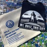 A navy blue T-shirt and a beige tote bag are laid out on a colorful background featuring cartoon illustrations of Everton Football Club fans, players, and banners. The T-shirt has a white graphic design showing a silhouette of Goodison Park's iconic tower and fence, with the text "WE BUILT THIS CITY ORIGINALS SINCE 1878" below it. The tote bag has a blue circular logo with a tower and the text "GOT" in the center, surrounded by a wreath. Below the logo, the bag reads "IT'S A GRAND OLD TEAM TO SUPPORT FOR EVERTONIANS, BY EVERTONIANS WWW.GRANDOLDTEAM.COM" in blue text, with a smaller note at the bottom saying "PLEASE KEEP THIS BAG OUT OF REACH OF BABIES AND SMALL CHILDREN." The background includes various Everton-related slogans and imagery, such as "EVERTON F.C. SINCE 1878" and "GOODISON PARK."