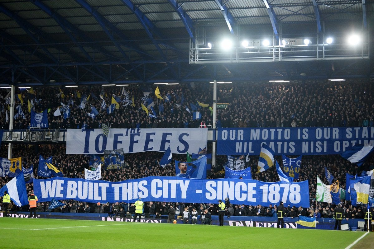 A prominent banner in the center reads, "WE BUILT THIS CITY - SINCE 1878," referring to Everton's founding year and their deep-rooted history in Liverpool. Another large banner to the left says "TOFFEES," a nickname for Everton FC, while a banner on the right displays the Latin phrase "NIL SATIS NISI OPTIMUM," which translates to "Nothing but the best is good enough," the club's motto. The stands are packed with supporters waving flags, including some with images of notable figures associated with the club, and the atmosphere appears electric under the stadium's bright floodlights. The field is visible in the foreground, with security personnel and other staff present near the sidelines.