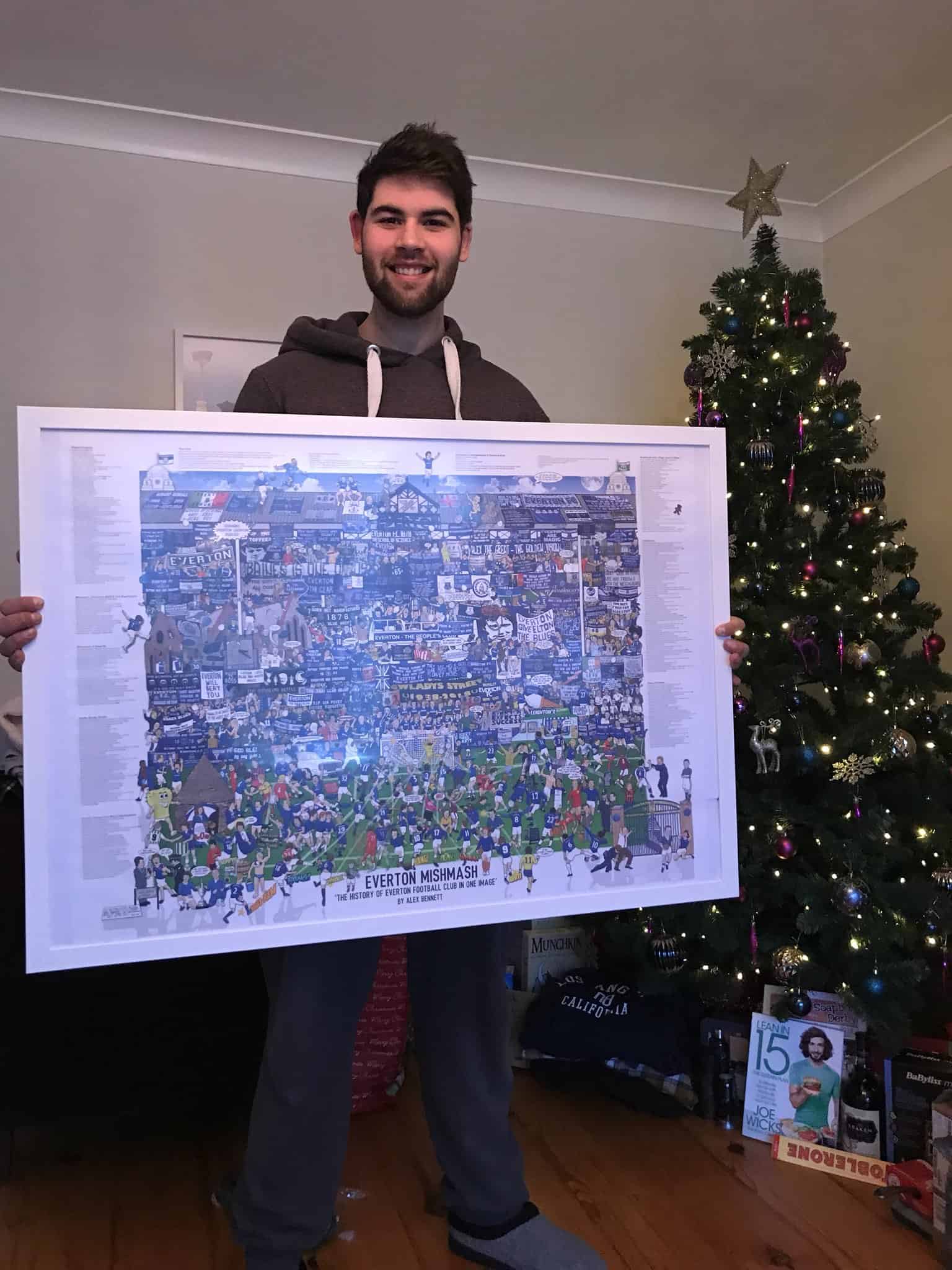 A smiling Toffees fan displaying his large framed Everton history poster, ideal inspiration for Everton gifts for dad.