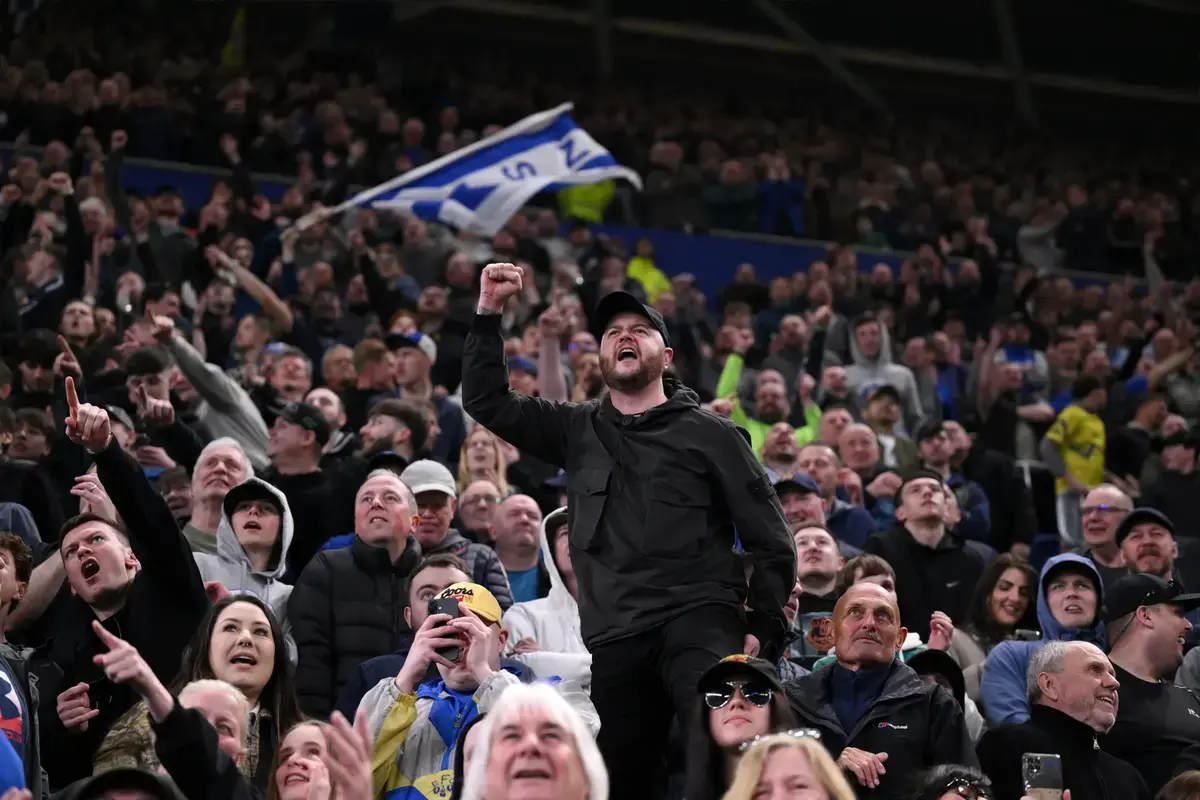 Everton fan singing passionately with raised fist surrounded by fellow supporters waving the Everton flag at the stadium