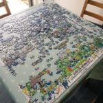 The image depicts a partially assembled jigsaw puzzle spread out on a light green tablecloth with a polka-dot pattern, placed on a dining table. The puzzle features a colorful illustration titled "Everton Mishmash: The History of Everton Football Club in One Image", showcasing various scenes, players, and elements related to Everton FC, with a dominant blue and green color scheme. The table is situated indoors near a glass door that opens to an outdoor patio area with potted plants and a pink watering can visible. Two wooden chairs are positioned at the table, and a cardboard box is seen in the background. The floor is covered with dark tiles, and natural light streams in from the patio area.