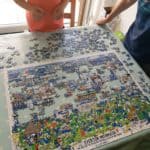 The image shows two individuals standing at a table, working on a partially assembled jigsaw puzzle. The puzzle depicts a colorful and detailed illustration titled "The History of Everton Football Club in One Image", featuring various scenes, players, and elements related to Everton FC. The puzzle pieces are spread out across a light-colored table with a polka-dot tablecloth, and some pieces are still unassembled. The setting appears to be indoors near a large window or glass door, with natural light illuminating the scene. The individuals are casually dressed, with one wearing a coral-colored shirt and the other a dark blue shirt with a striped pattern.