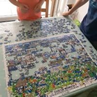 The image shows two individuals standing at a table, working on a partially assembled jigsaw puzzle. The puzzle depicts a colorful and detailed illustration titled "The History of Everton Football Club in One Image", featuring various scenes, players, and elements related to Everton FC. The puzzle pieces are spread out across a light-colored table with a polka-dot tablecloth, and some pieces are still unassembled. The setting appears to be indoors near a large window or glass door, with natural light illuminating the scene. The individuals are casually dressed, with one wearing a coral-colored shirt and the other a dark blue shirt with a striped pattern.