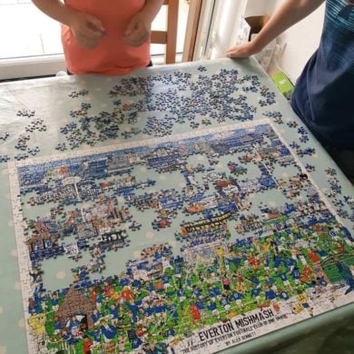 The image shows two individuals standing at a table, working on a partially assembled jigsaw puzzle. The puzzle depicts a colorful and detailed illustration titled "The History of Everton Football Club in One Image", featuring various scenes, players, and elements related to Everton FC. The puzzle pieces are spread out across a light-colored table with a polka-dot tablecloth, and some pieces are still unassembled. The setting appears to be indoors near a large window or glass door, with natural light illuminating the scene. The individuals are casually dressed, with one wearing a coral-colored shirt and the other a dark blue shirt with a striped pattern.